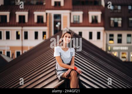 Una ragazza siede sul tetto di una casa in città la sera. Ritratto di un modello in un abito e sneakers Foto Stock