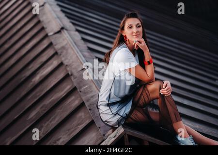 Una ragazza siede sul tetto di una casa in città la sera. Ritratto di un modello in un abito e sneakers Foto Stock