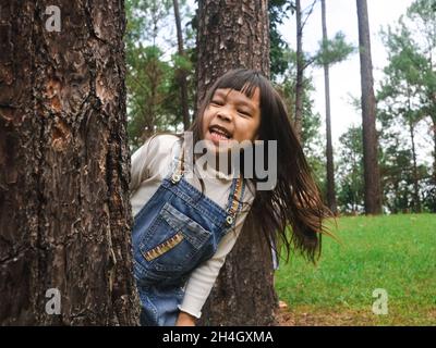 Carina bambina in piedi dietro un grande albero e sorridente alla macchina fotografica. Bambino felice che gioca all'aperto. La famiglia ha trascorso del tempo insieme. Foto Stock