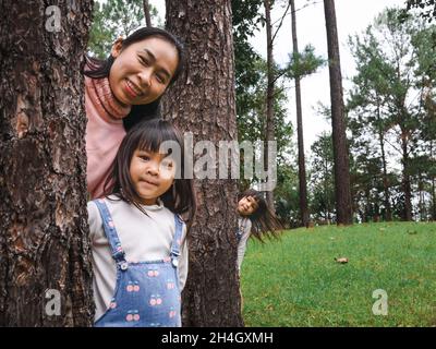 Felice madre e figlie in piedi dietro un grande albero e sorridendo alla macchina fotografica. Una famiglia felice che gioca all'aperto. La famiglia ha trascorso del tempo insieme. Foto Stock