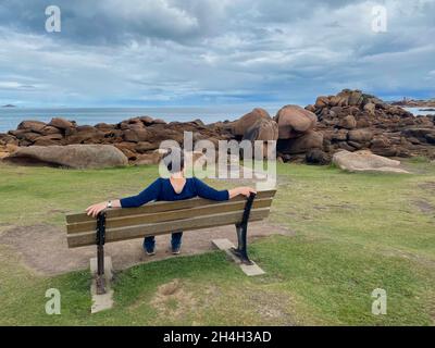 Donna anziana seduta su panchina, Ile Renote, costa rocciosa lungo il Sentier des douaniers, Tregastel, Cote de Granit Rose, Cotes d'Armor, Bretagna Foto Stock