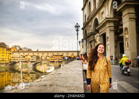 Una ragazza turistica con un cappotto d'arancio cammina felicemente sul lungofiume a Firenze, Italia. Foto Stock