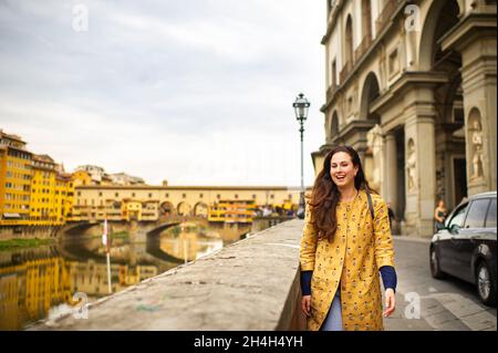 Una ragazza turistica con un cappotto d'arancio cammina felicemente sul lungofiume a Firenze, Italia. Foto Stock