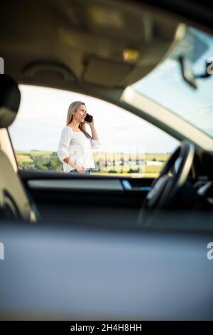 Donna piuttosto anziana al volante della sua auto, con una pausa durante un lungo viaggio, facendo una telefonata Foto Stock