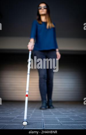 Giovane donna con visione compromessa che cammina per le strade della città, usando la sua canna bianca per navigare meglio lo spazio urbano e raggiungere la sua destinazione in modo sicuro Foto Stock