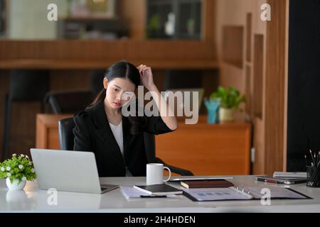 Dipendente femminile confuso con il suo lavoro su computer portatile in ufficio sul posto di lavoro. Donna d'affari pensiva in ufficio. Foto Stock