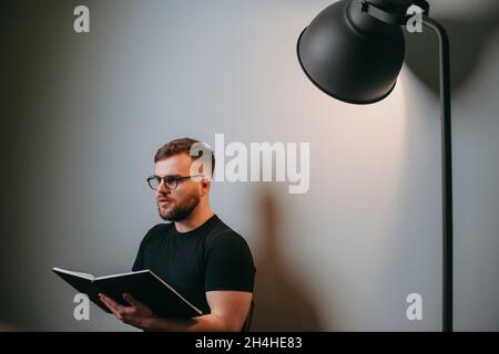 Uomo d'affari caucasico con barba in t-shirt nera seduta su sedia sullo sfondo di una parete bianca illuminata da una lampada. Vista laterale Foto Stock