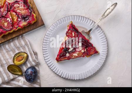 Pezzo di crostata di susina su piastra leggera con lama a spalla, accanto alla torta e prugne su tovagliolo. Vista dall'alto Foto Stock