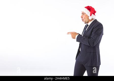Uomo d'affari vestito di vestito, cravatta e cappello di Natale di Babbo Natale, sorpreso, puntando con le mani, su sfondo bianco. Conc. Festa di Natale Foto Stock