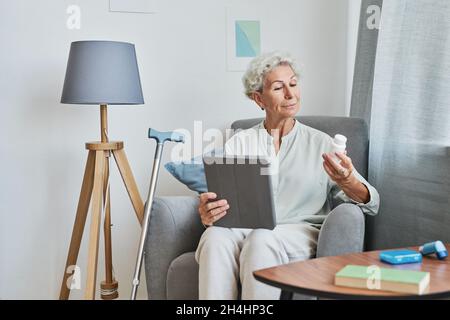 Ritratto di donna anziana moderna lettura flacone di prescrizione a casa e utilizzando tablet digitale, spazio copia Foto Stock