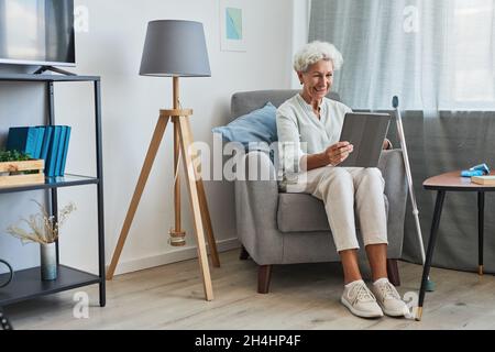 Ritratto a tutta lunghezza di una donna anziana sorridente utilizzando un tablet digitale a casa mentre si siede in una comoda poltrona con canna, spazio copia Foto Stock