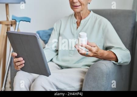 Primo piano della donna anziana moderna che tiene il flacone di prescrizione a casa e utilizzando il tablet digitale, spazio copia Foto Stock