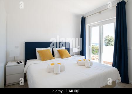 Camera matrimoniale con biancheria da letto bianca, testiera blu e porte scorrevoli in vetro per la terrazza Foto Stock