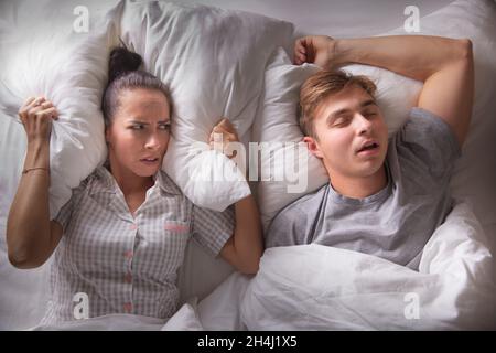 La donna guarda il suo ragazzo infastidito, tenendo il cuscino sopra le sue orecchie come russare steso sulla schiena con la bocca aperta. Foto Stock