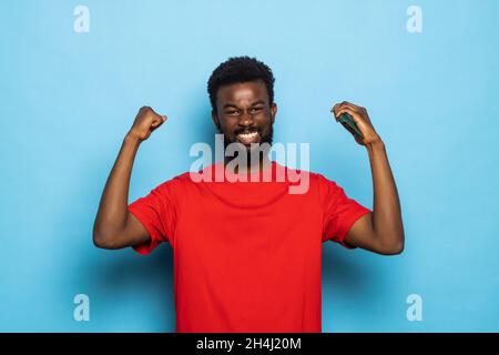 African American uomo con barba felice ed entusiasta che esprimono gesto vincente. Successo e celebrando la vittoria, trionfante isolate su backgr blu Foto Stock