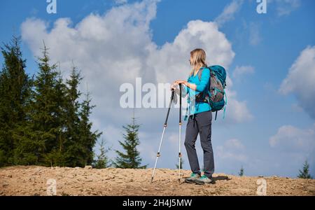 Giovane viaggiatore donna con zaino e racchette da trekking in piedi sul bordo della strada in pietra. Donna escursionista guardando il bel pino verde vicino a lei sullo sfondo di nuvole bianche su cielo blu. Foto Stock