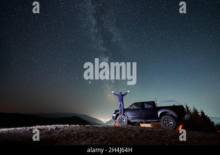 Vista posteriore dell'uomo con le braccia distese in piedi su una ruota anteriore in gomma di jeep nera nel mezzo di una strada di montagna rocciosa e guardando il cielo stellato fiabesco notte. La Via Lattea sulle montagne. Foto Stock