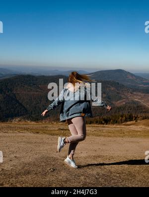 Una giovane donna in un vestito caldo e una giacca in denim gioisce in montagna. Autunno in montagna. Ragazza felice. Natura e viaggi. Foto verticale. Foto Stock