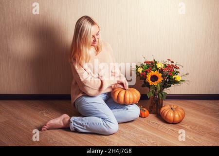 Giovane donna bionda in caldo maglione è seduta sul pavimento e tenere la zucca nelle sue mani e guardare. Foto Stock