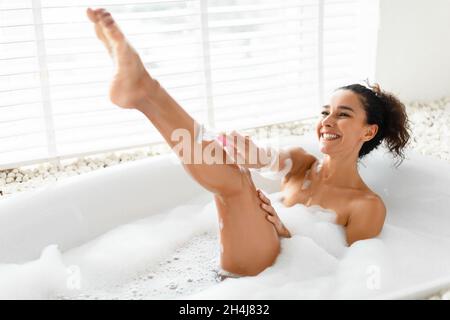 Bella giovane donna rasando le gambe con rasoio, facendo la procedura di rimozione dei capelli mentre prendendo bagno bolla a casa Foto Stock