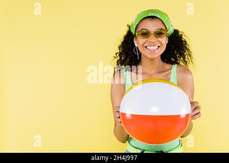 giovane donna afro-americana positiva in fazzoletto che tiene la palla gonfiabile isolato su giallo Foto Stock