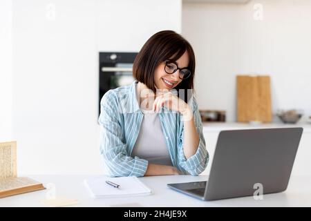 Concetto di lavoro freelance. Felice signora giovane seduta al tavolo con il computer portatile in cucina, guardando lo schermo e sorridendo Foto Stock