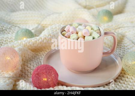 Coppa rosa di cacao con marshmallow su tele di maglia bianca con ghirlande leggere. Concetto di vacanze invernali. Primo piano. Foto Stock