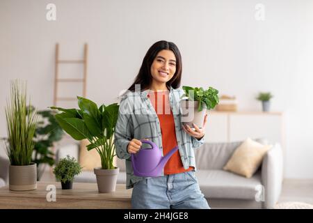 Ritratto di giovane donna araba sorridente con annaffiatoio che si prende cura di piante domestiche in casa Foto Stock