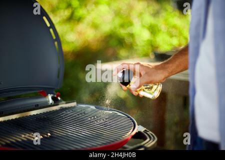 Primo piano sulla mano dell'uomo spruzzando olio su barbecue gas grill all'aperto nel cortile, preparazione per tostatura su Grill, picnic in famiglia estate, cibo su th Foto Stock