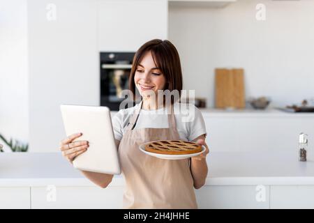 Buona giovane casalinga che mostra torta fatta in casa, videochiamata su tablet digitale, cucina in cucina moderna interno. Foto Stock