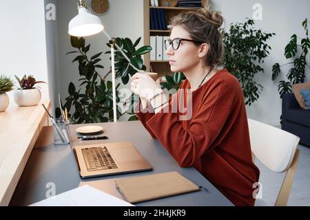 Giovane donna in occhiali seduti alla scrivania con un computer portatile e pensando al suo lavoro online a casa Foto Stock