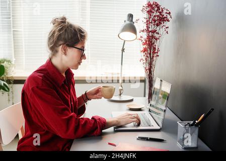 Giovane donna d'affari in occhiali seduti al tavolo bevendo caffè e lavorando online sul portatile a casa Foto Stock