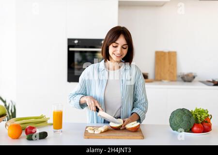 Felice giovane signora tagliare le mele fresche su asse di legno mentre fare Macedonia di frutta per la prima colazione in cucina interna, preparando pasto sano Foto Stock