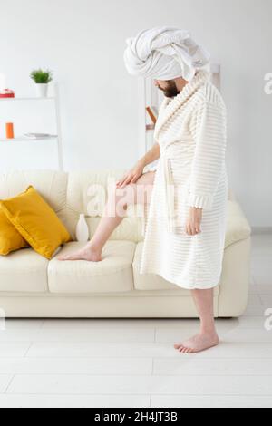 L'uomo bearded divertente sfrega la crema del corpo sulle sue gambe. Spa, cura del corpo e della pelle per l'uomo Foto Stock