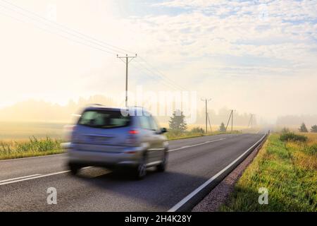 Auto che guida a velocità su un'autostrada rurale su una bella nebbia più tardi mattina estate. Sfocatura del movimento all'interno della fotocamera. Foto Stock