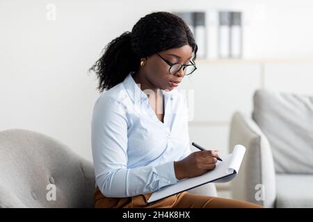 Ritratto di focalizzato psicologa femminile nera scrittura in appunti, seduta in poltrona in ufficio moderno. Psicoterapeuta sicura che prende appunti durina Foto Stock