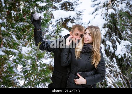 Coppia felice di uomini e donne all'aperto decorare albero di natale all'aperto in abiti caldi nel parco nevoso in inverno. Un ragazzo e una ragazza di aspetto caucasico abbracciano un umore festivo e romantico Foto Stock