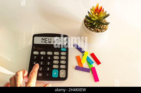 Calcolatrice con il testo New Life sul tavolo da ufficio con adesivi, fiore di cactus. Una mano femminile su sfondo bianco giace su una calcolatrice. Foto Stock