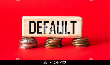 La parola DEFAULT è scritta su un blocco di legno che si trova su monete e uno sfondo rosso. Concetto di aumento del debito. Foto Stock