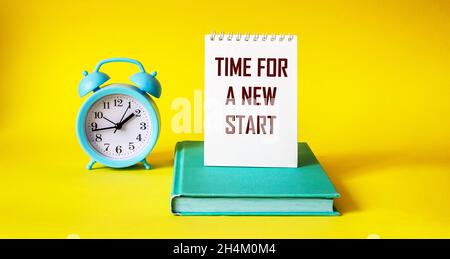 ORA DI UN NUOVO INIZIO, il testo è scritto su un notebook che si trova su un libro, accanto a una sveglia su sfondo giallo Foto Stock