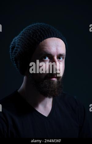 autentico ritratto drammatico di un millenario con una barba in un cappello di maglia nero, su sfondo scuro. Foto Stock