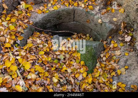 Un vecchio pozzo con un muro di pietre danneggiato è coperto di foglie autunnali Foto Stock