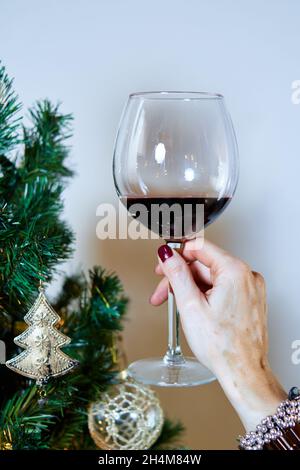 Le mani di una giovane donna con vitiligo tostano con un bicchiere di vino accanto all'albero di Natale decorato con ornamenti dorati. Foto Stock