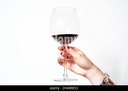 mano di una giovane donna con vitiligo e unghie dipinte di rosso tostando con un bicchiere di vino rosso su sfondo bianco. Foto Stock