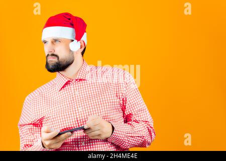 Un uomo portatore pensivo in un cappello di Natale di Babbo Natale tiene un telefono nelle sue mani Foto Stock