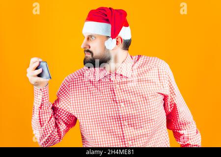 L'uomo bearded in una camicia di plaid ed il cappello di Babbo Natale è scioccato e guarda il telefono con sorpresa Foto Stock