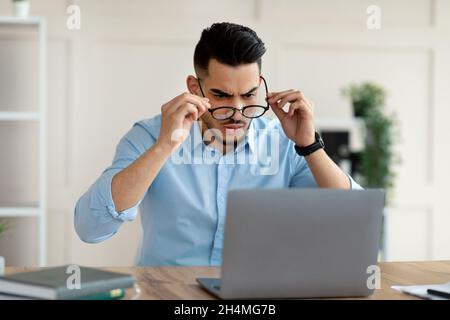 Irritato uomo d'affari arabo che guarda lo schermo del laptop, avendo problemi nel lavoro in linea in ufficio. Giovane dipendente aziendale con computer portatile feeling st Foto Stock