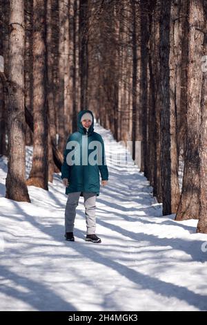 Donna caucasica che cammina nella foresta. Giovane donna attraente in giacca calda giù si trova su strada coperta di neve dopo la Blizzard e gode il sole su ghiaccio Foto Stock