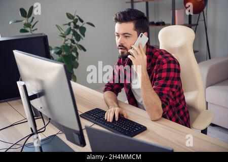 Foto di serio messo a fuoco giovane uomo scrivere computer programmatore tenere telefono parlare al chiuso all'interno del posto di lavoro ufficio Foto Stock