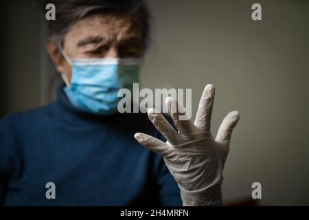 Guanto medico bianco in primo piano, mostrato da una nonna anziana che indossa una maschera medica, chiedendo sicurezza e igiene durante un'epidemia Foto Stock